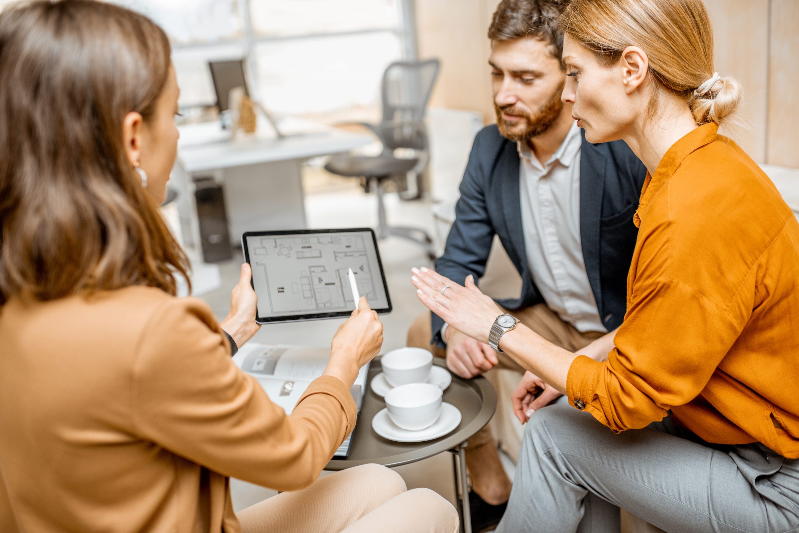 Image représentant une femme expert comptable avec une chemise marron en train de montrer une tablette avec des informations à un couple autour d'une tasse de café